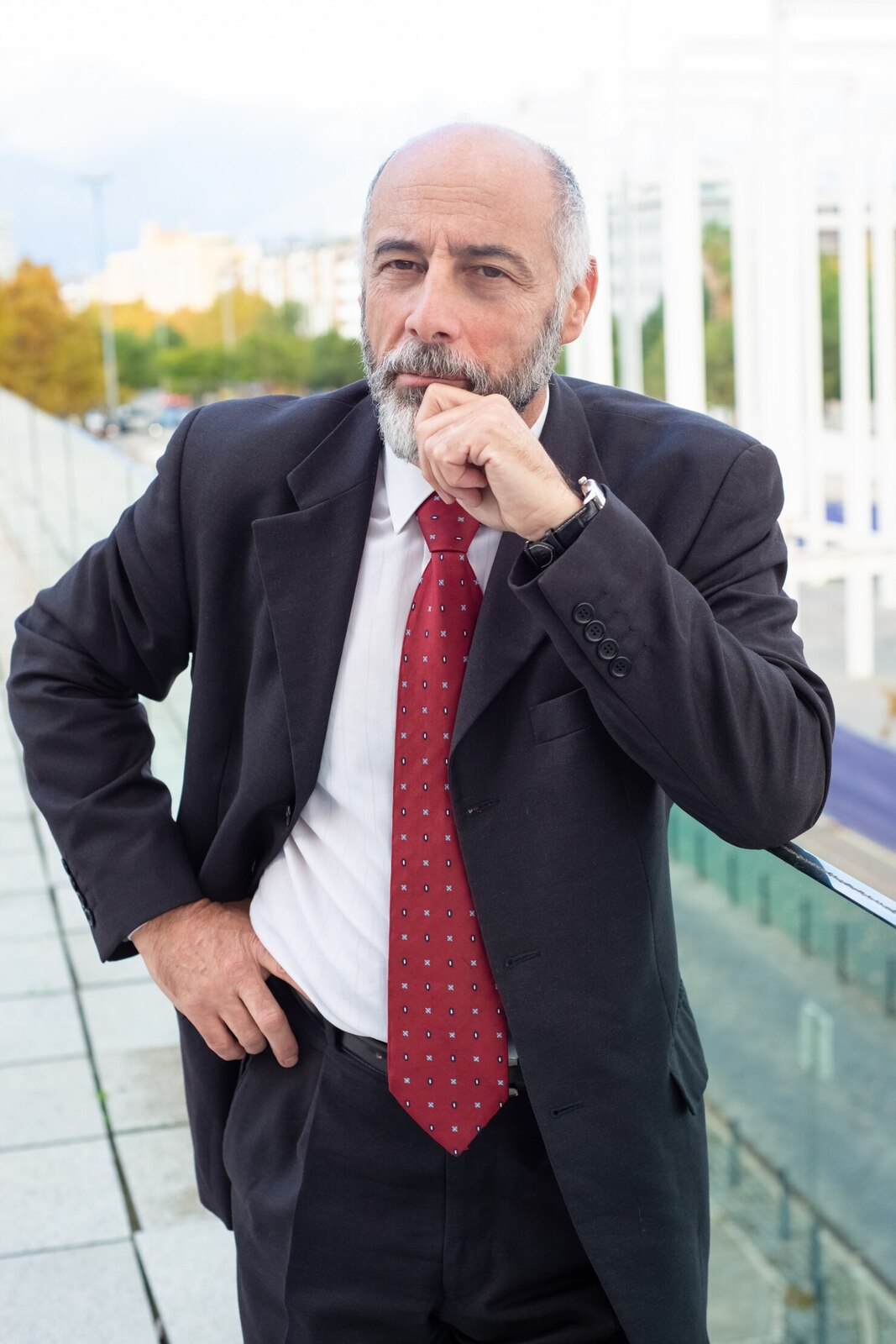 A smiling man with gray hair, glasses, and a beard, wearing a dark pinstripe suit, white shirt, and blue tie, standing against a plain background.